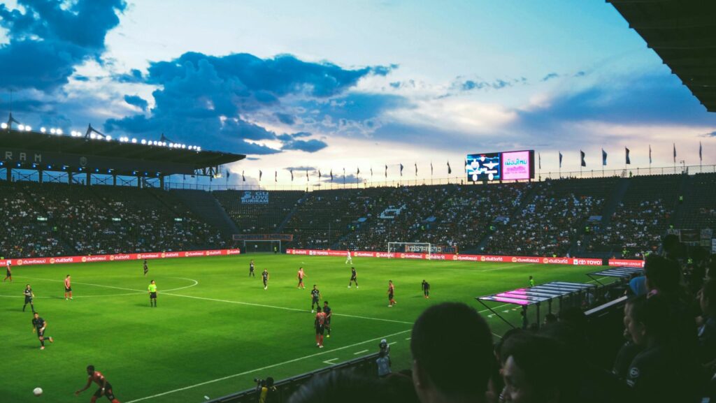 A football match in progress at a large stadium with spectators and a sunset sky